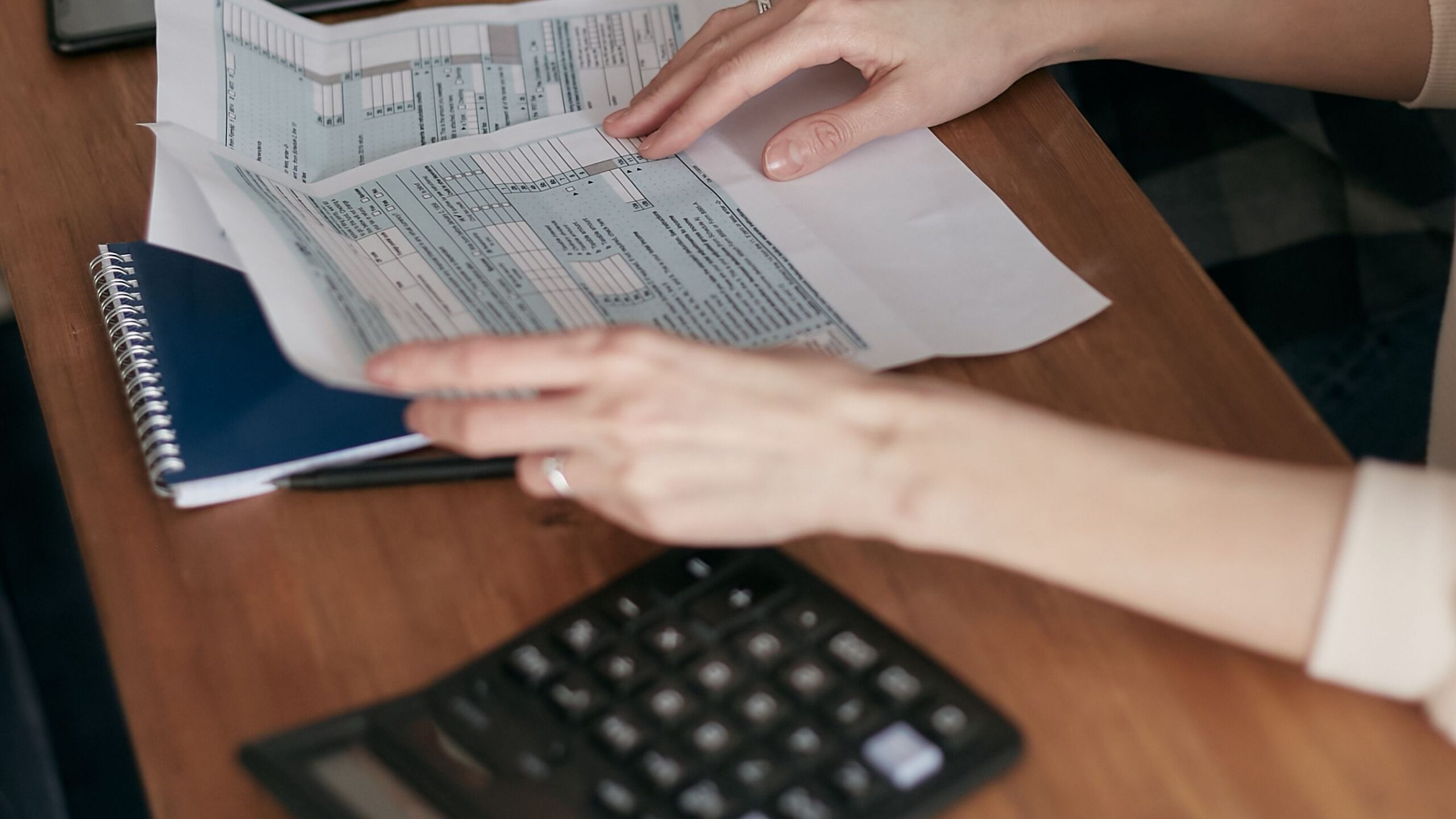 Close-up of hands organizing tax forms on a desk with a calculator, laptop, and notebook.