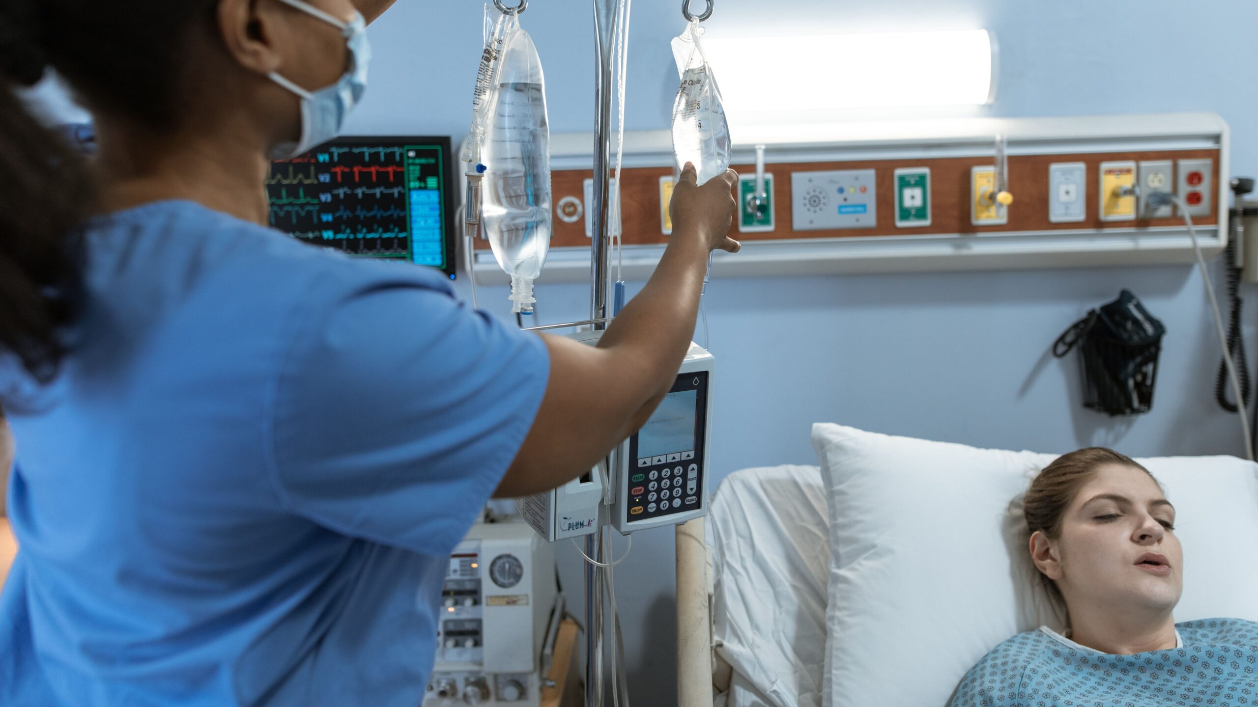 A nurse in a hospital setting adjusts an IV drip for a patient lying in bed, showcasing medical care.
