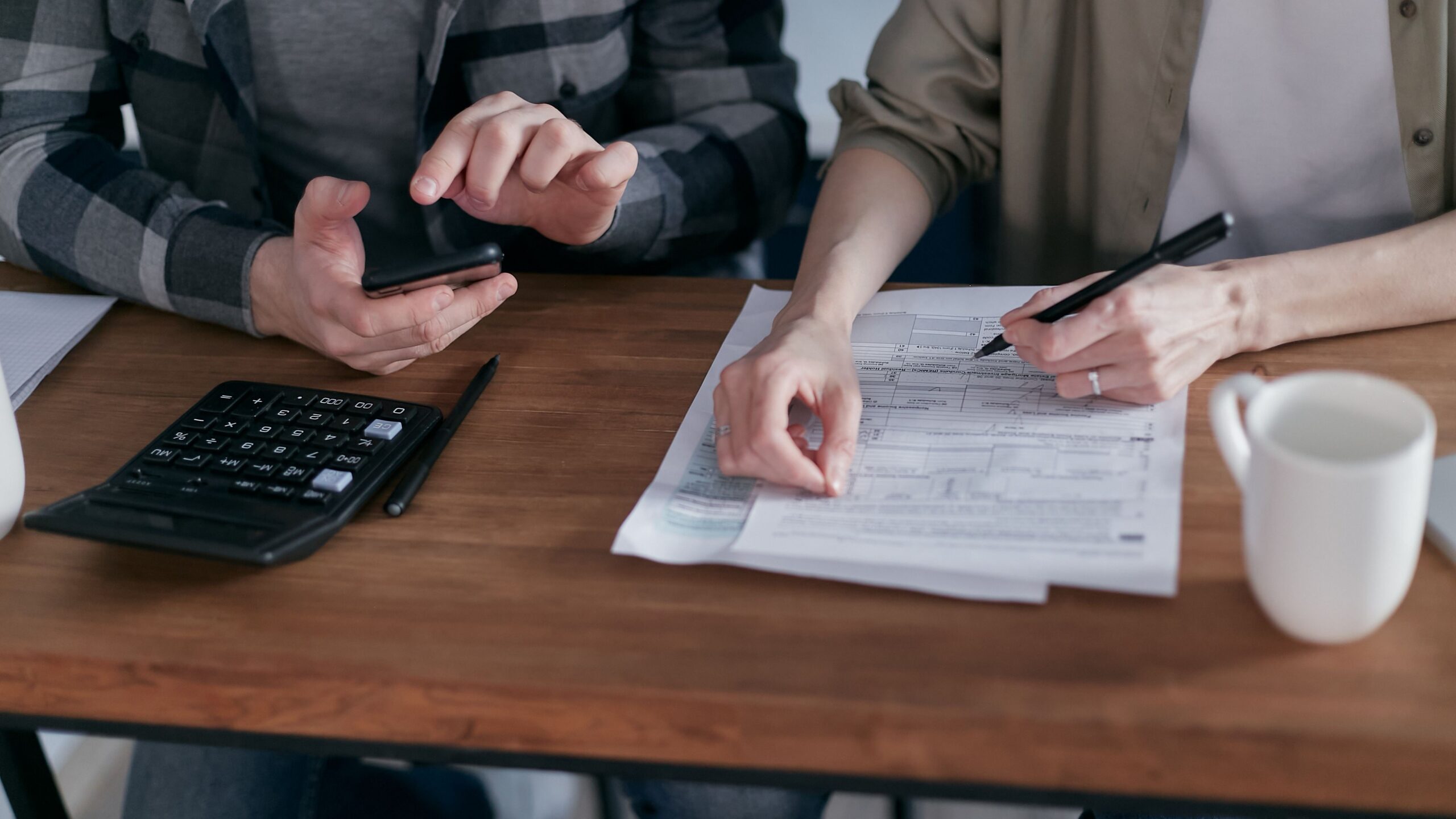 Two people working together on tax forms using a calculator at a wooden desk.