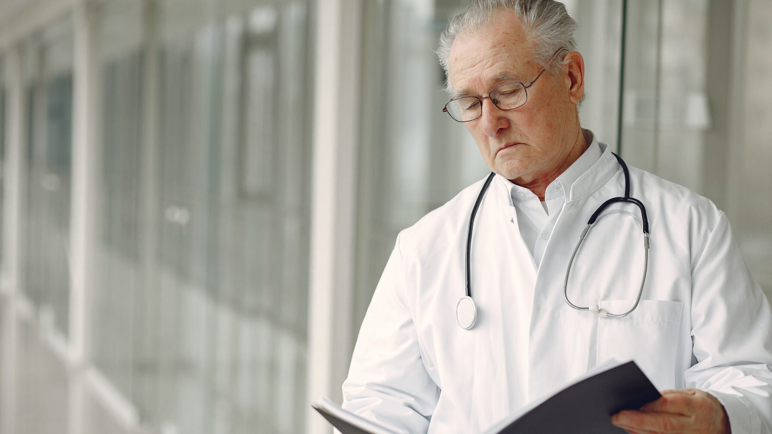 Crop concentrated professional senior male physician in white robe with stethoscope standing in modern hospital hallway and reading medical case records in daylight