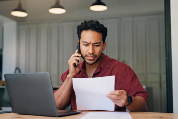 Laptop documents and businessman with phone call in office with contact for programming mistake Computer confused and web developer on mobile discussion for feedback on coding error with paperwork