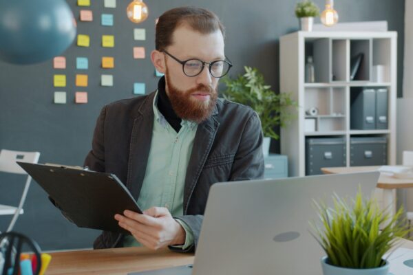 Man with beard working on laptop and clipboard