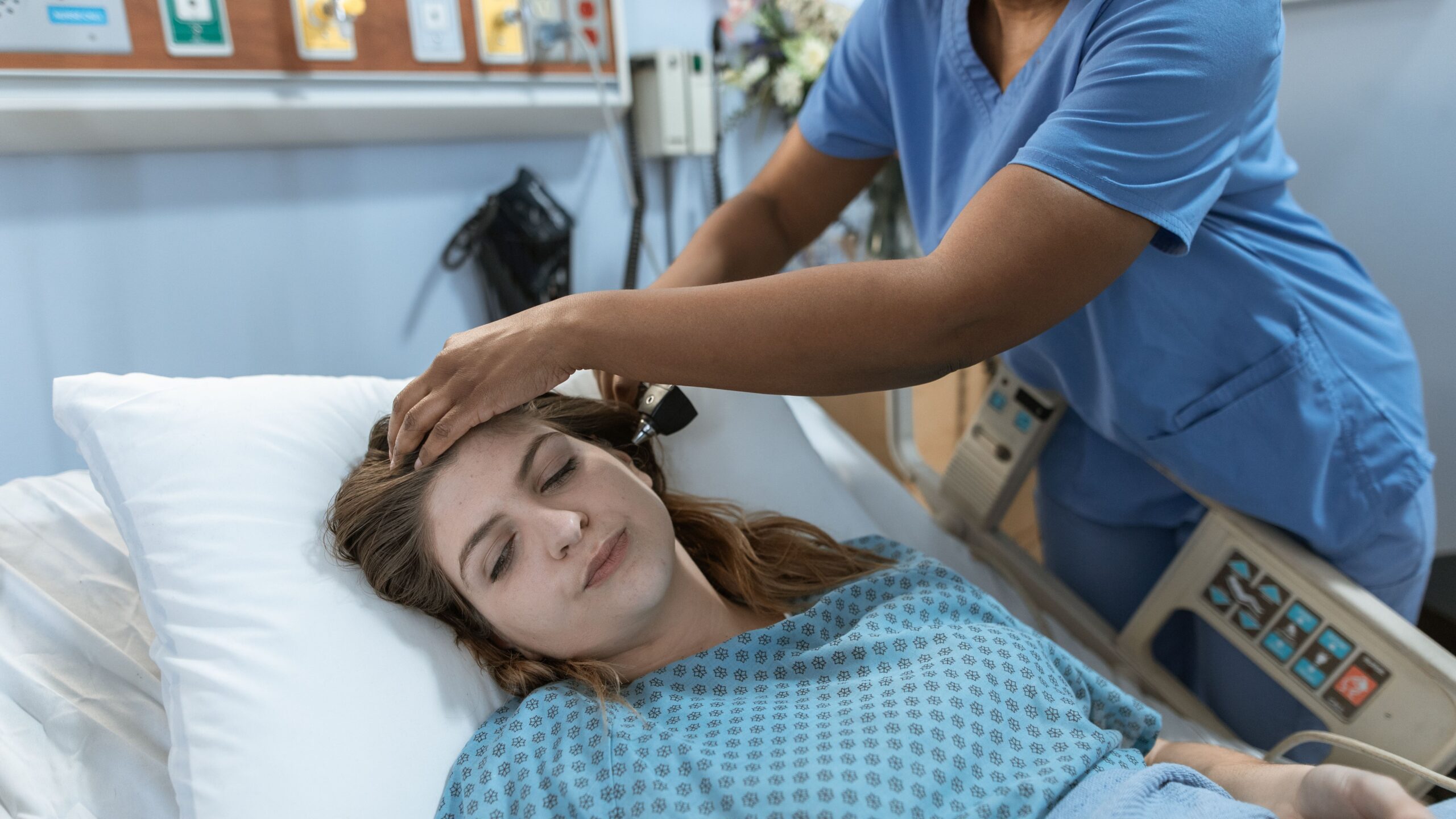 Crop African American female doctor with professional equipment doing examination of ear of woman lying on bed in hospital ward
