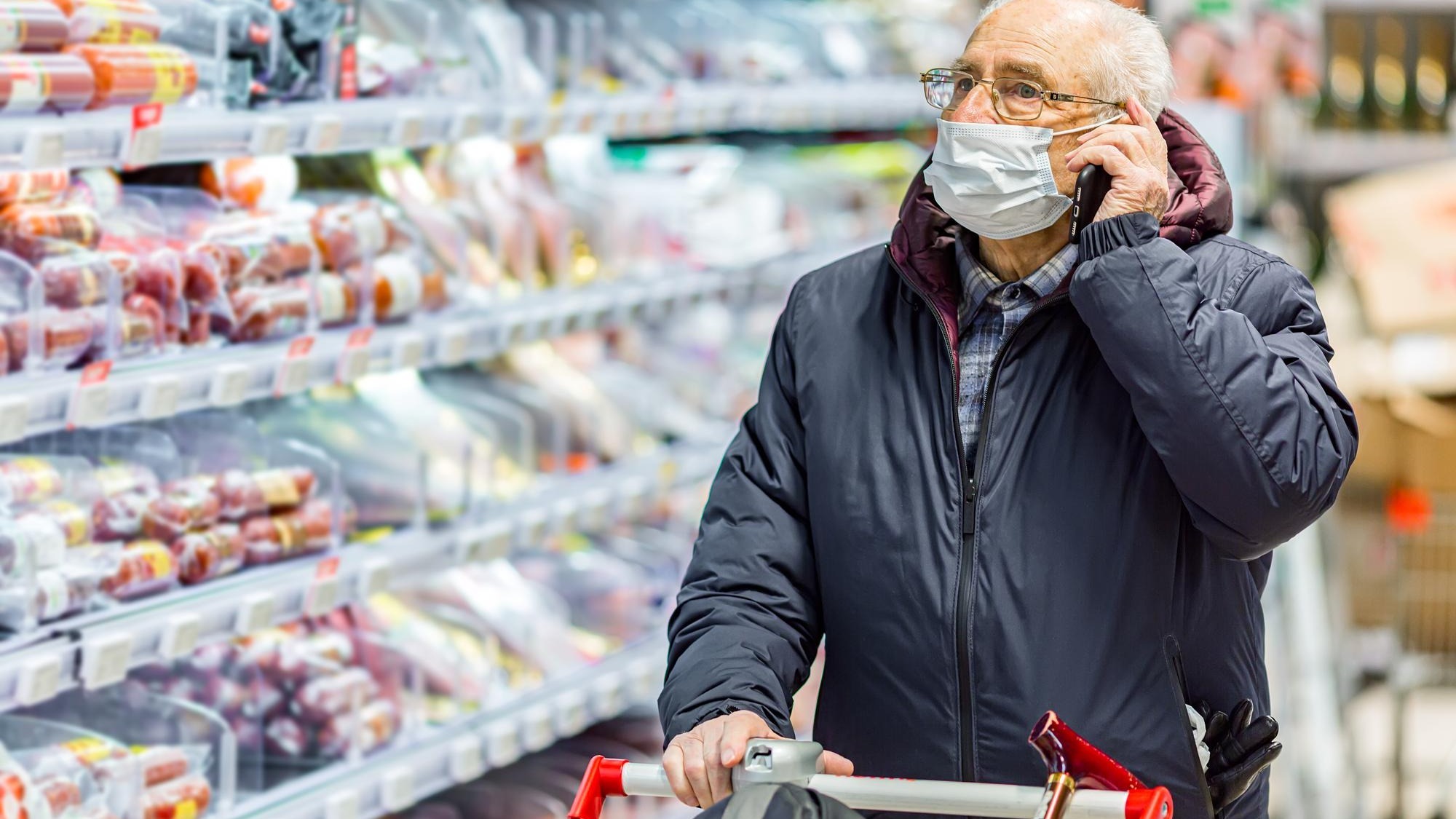 Old senior european man wearing protective facial mask pushing shopping cart and talking on the phone in the supermarket. Shopping during COVID-19 concept.