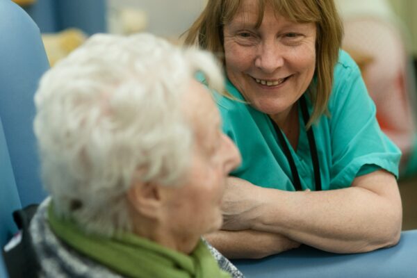 A smiling woman in green scrubs leaning toward an elderly person in a chair