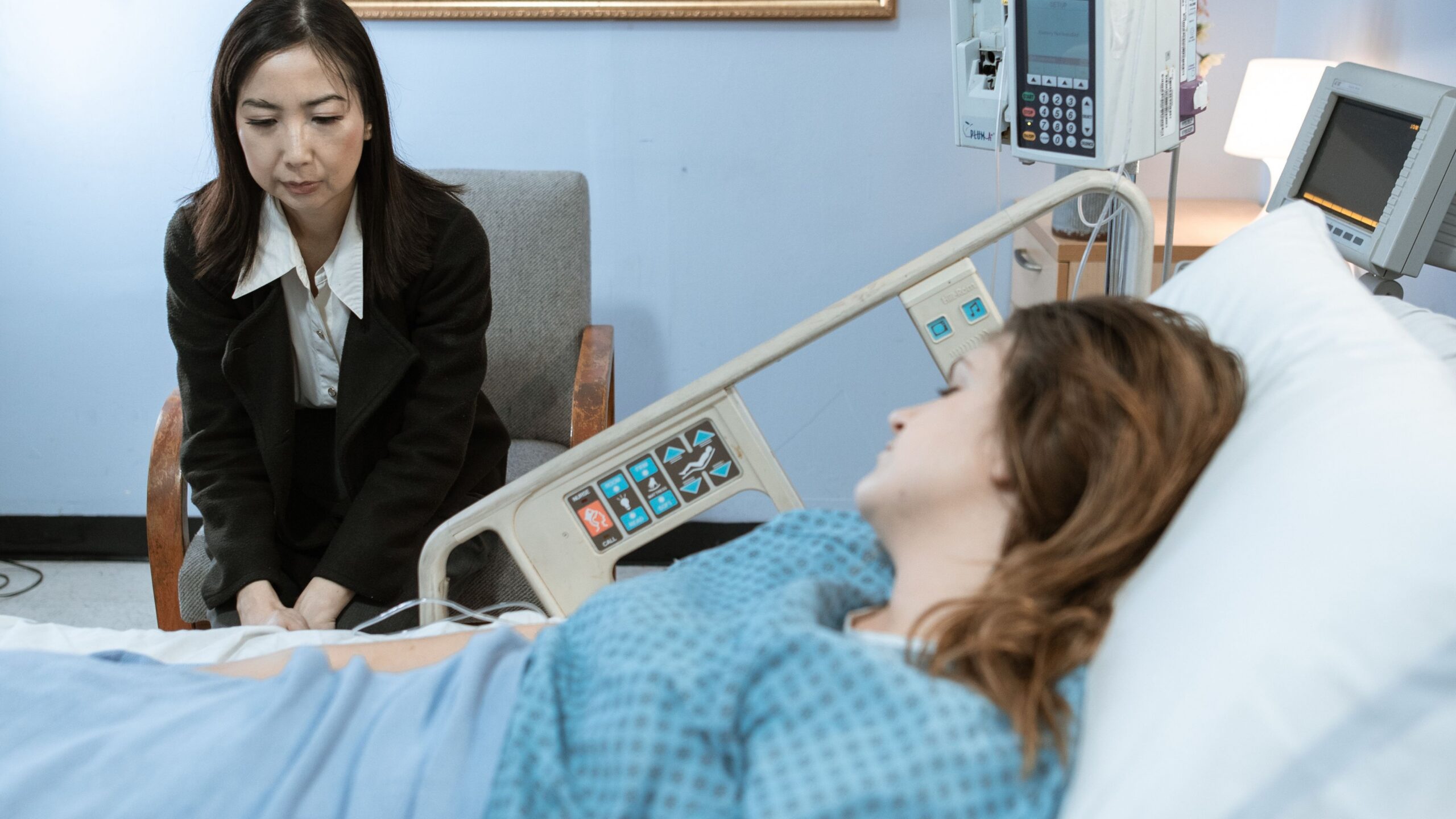 A hospital room showing a patient in bed and a visitor sitting beside her.