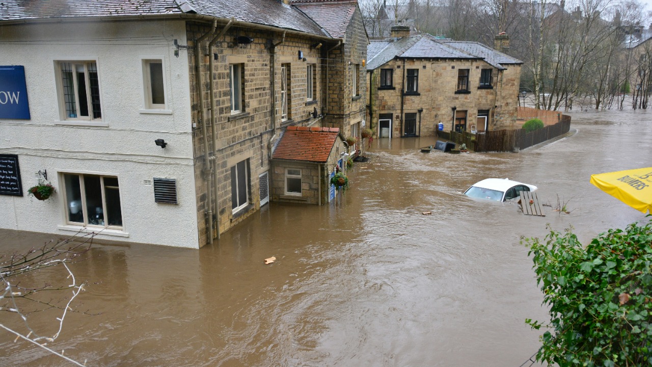 Floodwaters surrounding stone buildings and a partially submerged white car in a town