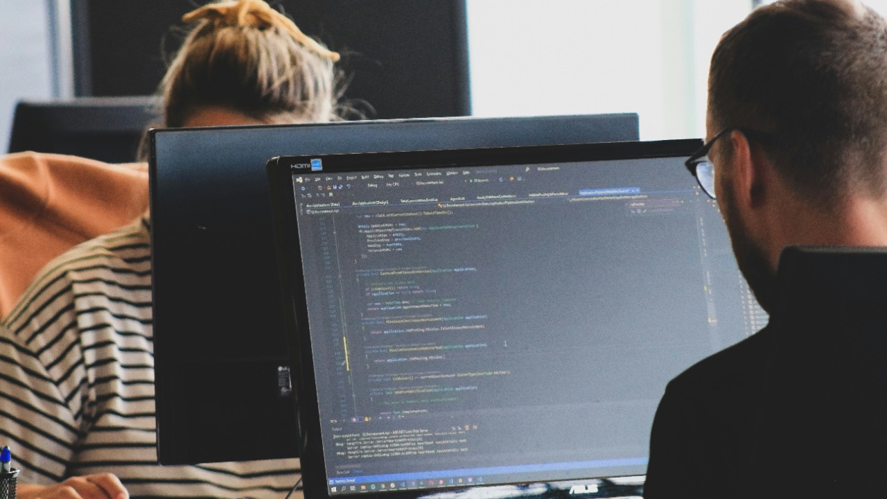 Two people working on computer code at monitors in a bright office workspace