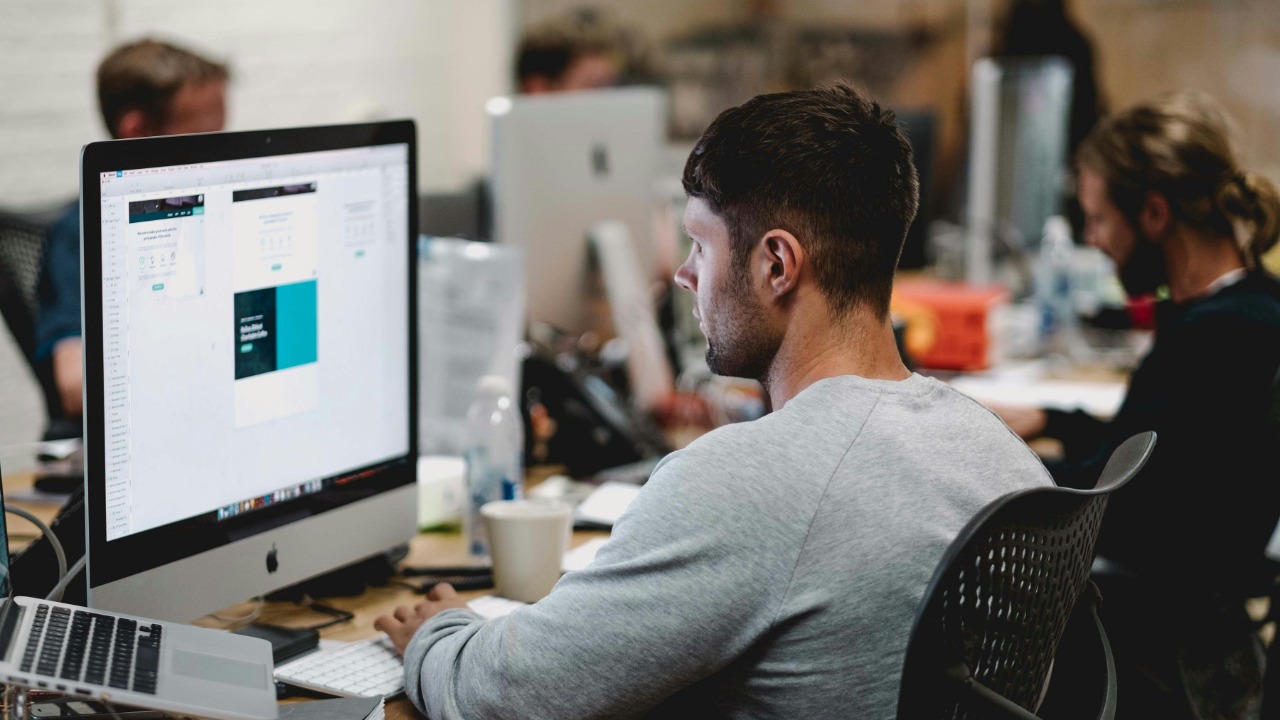 man in gray sweatshirt sitting on chair in front of iMac
