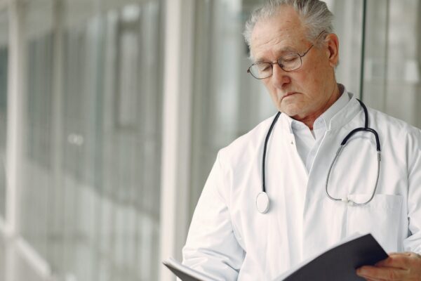 Crop concentrated professional senior male physician in white robe with stethoscope standing in modern hospital hallway and reading medical case records in daylight