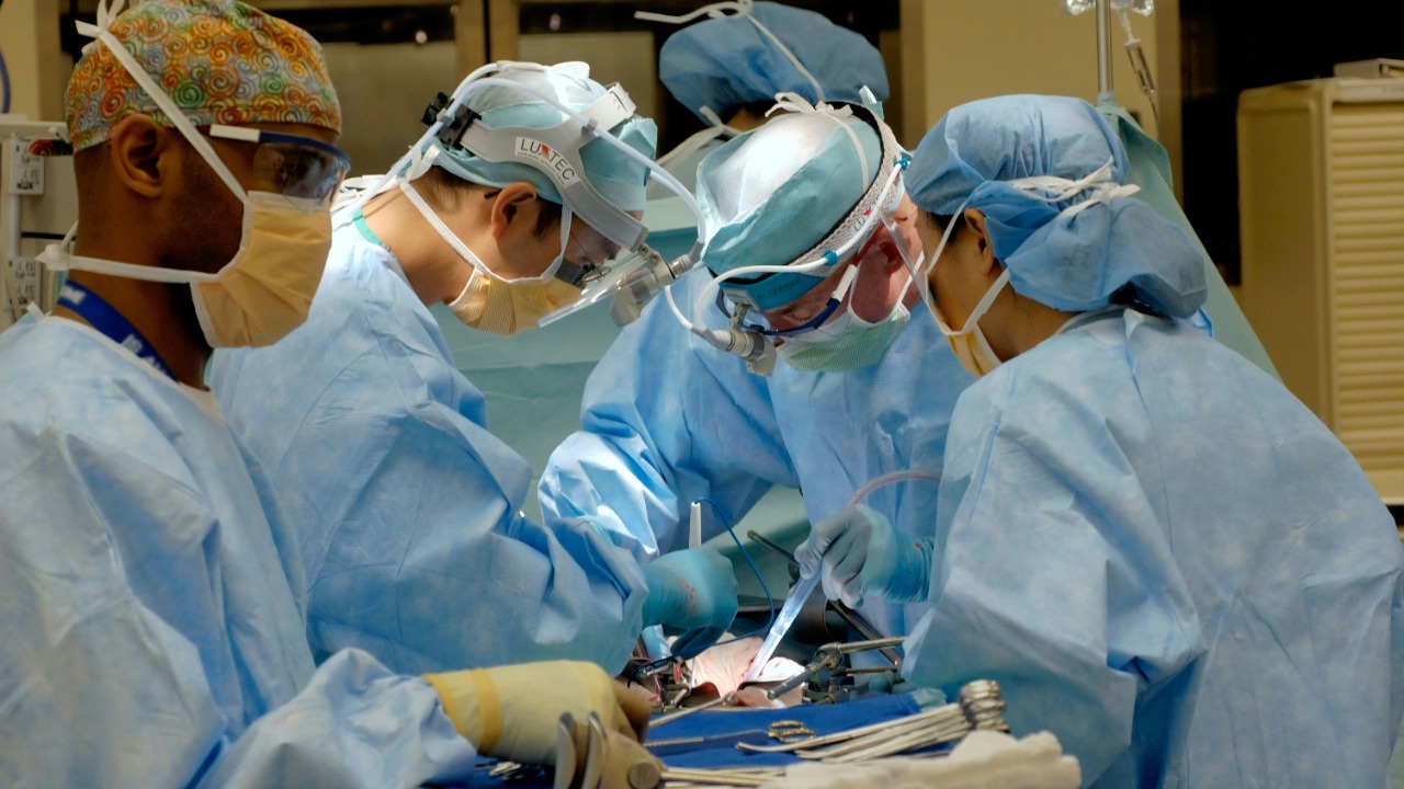 Four surgeons in blue scrubs and masks performing surgery in a bright operating room
