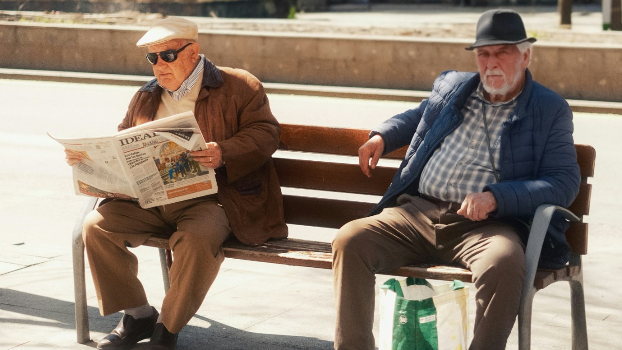 Two elderly men sitting on a park bench reading newspaper