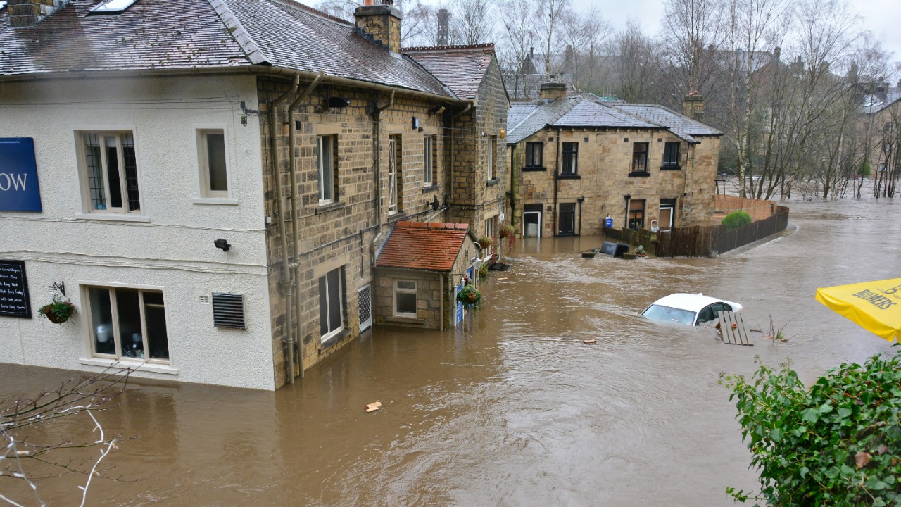 Floodwaters surrounding stone buildings and a partially submerged white car in a town